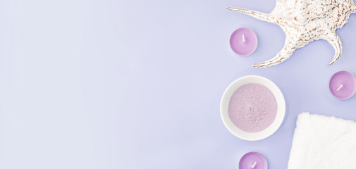 Cosmetic clay on lilac pink background, next to a towel, sea shell and with candles. The concept banner of beauty cosmetic procedures, spa, skin treatment and skin care. Copy space