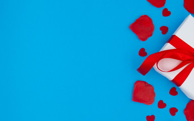 Red rose petals and red hearts lying on a blue background. Gift wrapped in white paper and a red ribbon.