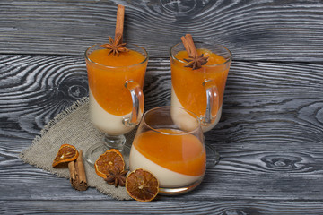 Two layers of jelly in glass goblets. Cream and tangerine jelly. In jelly, cinnamon stick and anise. Near spices for decoration. On the surface of brushed boards painted black and green.