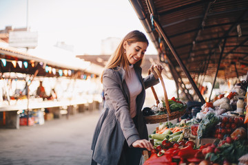 Young cheerful woman choosing vegetables and holding her basket at farmer's market.
