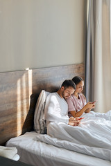 Obraz na płótnie Canvas Young pleasant lovers in pajamas having happy time in hotel room, sun shining through big window, texting with friends using cell phones, looking away with happy faces,relationship and travel concept