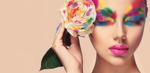 Fototapeta premium Beautiful girl model with multi-colored paints on her face. Woman with rose flower and bright color make-up. Cosmetics, beauty and makeup. Spring and summer flowering shopping