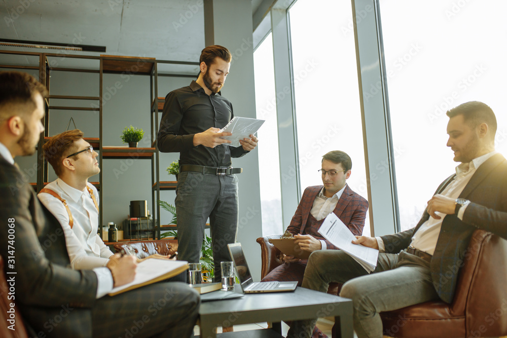 Wall mural serious speaker man in black formal shirt stand giving speech to co-workers, holding paper document in hands. isolated in modern boardroom with panoramic window