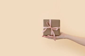 Female hand holding gift box wrapped in craft paper with pink ribbon through on pale beige...