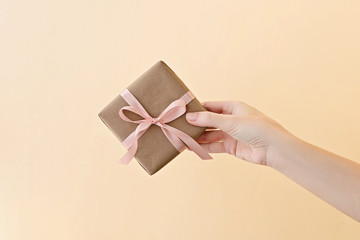 Female hand holding gift box wrapped in craft paper with pink ribbon through on pale beige background.