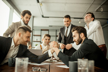 positive caucasian co-workers enjoy free, leisure time at work place, two men play arm wrestling in office