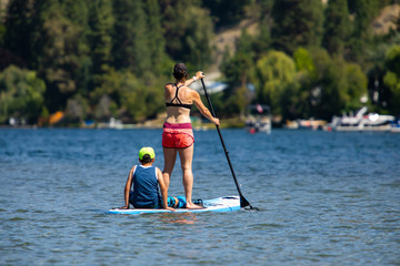 Back view of a young woman with son paddle boarding in sports clothing. Wide shot of mom and son during active holidays in sunny day