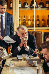 Thoughtful bearded businessman resting elbows on light wooden table attentively listening to presentation of young partner looking aside