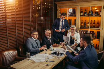 Bored businessman in glasses wearing gray stylish suit sitting thoughtfully at meeting with partners, holding pen in hands posing against background of jalouse, side view