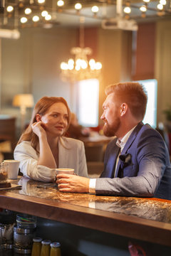 Close up of couple having first date in romantic place. Beautiful young woman looking at boy friend with admire, lovely man holds a cup of coffee, turns head towards pretty girl with kind smile