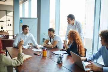 A team working on a business project actively. 6 people at the office having conference, discussing work, researching and creating charts to calculate the best strategy. Colleagues brainstorming.