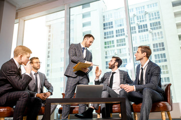 young caucasian business colleagues sit on leather chairs and discuss together in modern office, wearing elegant suits. big panoramic window in the background