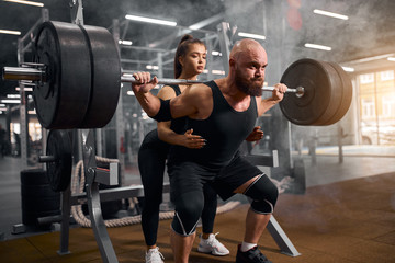 Fototapeta na wymiar Supportive young pretty female trainer standing behind brutal hairless man, squatting with heavy barbell in brightly lighted fitness room, demonstrates right position, fitness performing