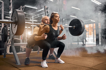Fototapeta na wymiar Cool attractive sporty fitness woman dressed in black sportswear, squatting using heavy barbell, taking help of professional fitness trainer, training in modern gym, preparing for competition