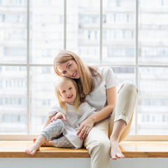 Obraz premium Portrait of young mother embracing her pretty daughter while sitting in front of window