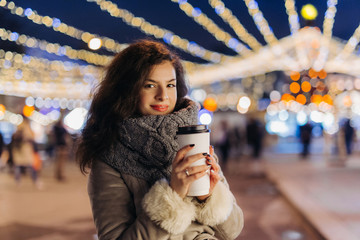 Girl Walking with a hot drink on christmas decoration lights street. Young happy smilin Woman...