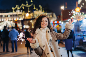 Girl having fun with sparklers on christmas decoration lights street. Young happy smilin Woman wearing stylish knitted scarf Outdoors. Model laughing. Winter wonderland city  scene New Year party