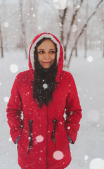 Portrait of young woman in red jacket and hood in winter season. Beautiful lady smiles, rejoicing in fall of snow. Fluffy snow envelops everything around.