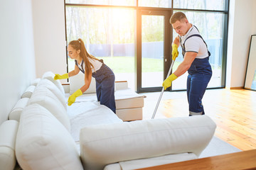 Two janitors in uniform cleaning new room with white sofa, mopping floor, straighten sofa. Work in...