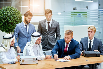 Caucasian man gice document for sign. Caucasian men in tuxedo, sheikhs in white suit. Office background