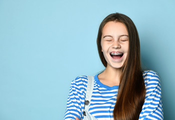 Teenager in jeans overall and striped sweatshirt. She is laughing with closed eyes, posing on blue studio background. Close up