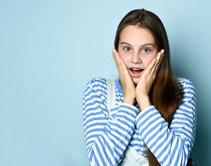 Teen lady in jeans overall and striped sweatshirt. She is looking surprised while posing on blue studio background. Close up