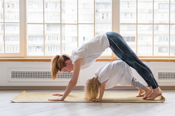 Young mother doing yoga with 3-years girl in front of window. Downward facing dog asana pose