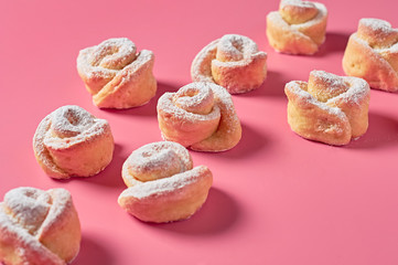 Scattered many homemade baked buns in form of rose spangled of powdered sugar lies on pink background. Close-up