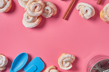 Heap of many homemade baked buns in form of rose spangled of powdered sugar near metal stencils, spatula, brush, spoon and cinnamon sticks lies on pink desk. Confectioners workplace. Flat lay