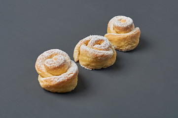 Row of three homemade baked buns in form of rose spangled of powdered sugar lies on dark concrete desk. Confectioners workplace. Close-up