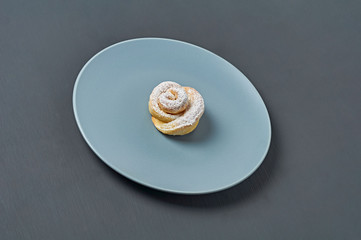 One homemade baked bun in form of rose spangled of powdered sugar on gray round plate lies on dark concrete desk. Confectioners workplace. Close-up