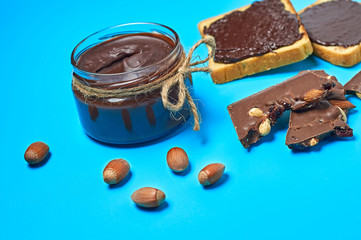 Single glass transparent jar with rope full of melted chocolate or cream near two pieces of vanilla bread and scattered hazelnut lies on blue desk on kitchen. Concept of nutritious breakfast or lunch