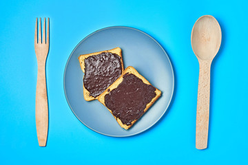 Two pieces of vanilla bread with melted chocolate on gray plate near wooden spoon and fork lies on blue desk on kitchen. Concept of nutritious breakfast or lunch