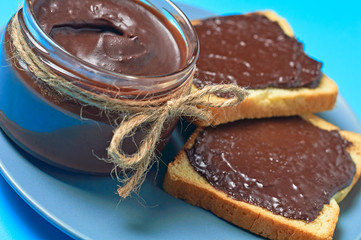 Glass jar full of melted chocolate on gray plate near pieces of vanilla bread lies on blue desk on kitchen. Concept of nutritious breakfast or lunch