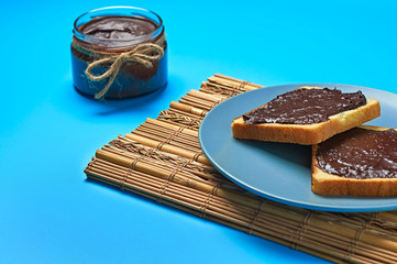 Glass jar with melted chocolate or cream near pieces of vanilla bread on plate and bamboo mat lies on blue desk on kitchen. Concept of nutritious breakfast or lunch