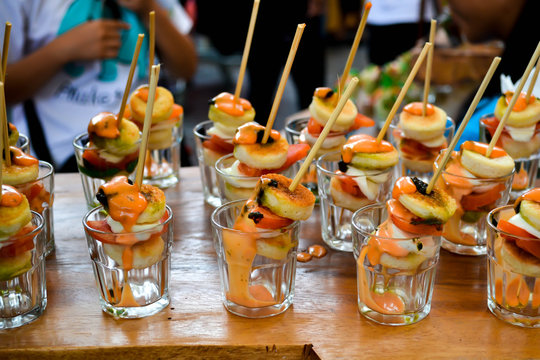 Snacks Set On Wooden Table. People In The Background Tasting Street Food. Seafood Skewers With Dressing In Glass Shots. Delicious Luxury Appetizer. Degustation, Cooking Festival, Market. Close Up.