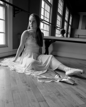 B&W photo of pretty ballet dancer sitting in front of window putting on pointe shoes.