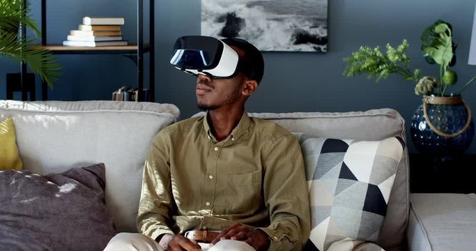 African American young guy in VR glasses sitting on the sofa in the cozy living room and having virtual reality headset at home. Indoor.