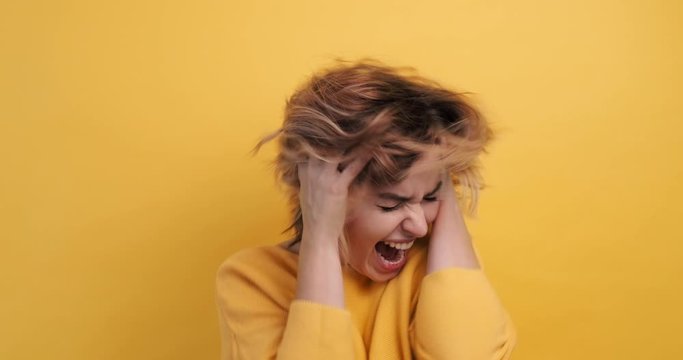 Slow motion woman screaming hysterics clutching both hands over head with frightened face, looking directly camera, isolated yellow background. Depression, fright, angry. Negative emotion. Monotone