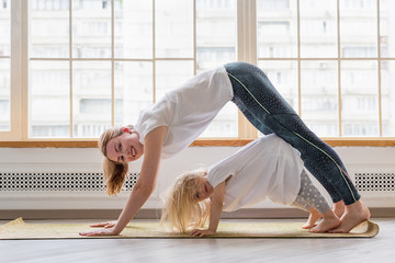 Young mother doing yoga with 3-years daughter in front of window. Happy mom smiling while...