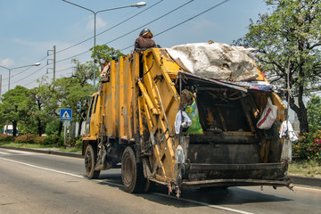 The garbage man sits on a garbage truck driving down Bangkok street.