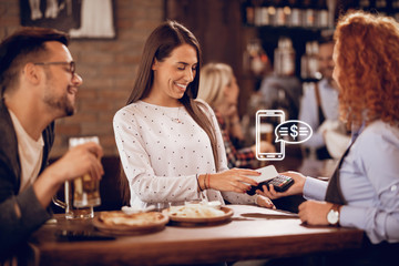 Happy woman making contactless payment to a waitress in a bar.