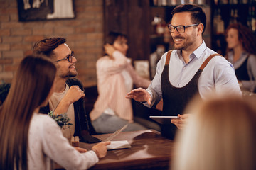 Happy waiter with touchpad talking to a couple in a pub.