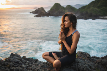 Young woman praying and meditating alone at sunset with beautiful ocean and mountain view....