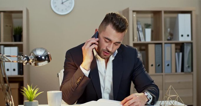 Caucasian businessman writing notes in the notebook and then answering a phone call while sitting at the table in his office.