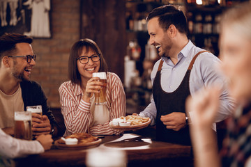 Happy waiter having fun with customers while serving them in a pub.