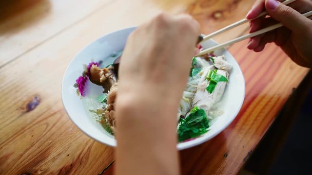 Woman's hands holding chopsticks and putting chicken slices and noodles to a Thai spoon. Woman having Thai chicken soup with rice noodles and greens for lunch at a street food cafe outdoors