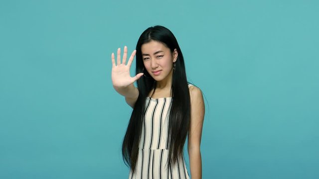 Angry bossy asian woman with long straight black hair in dress showing ban stop sign with raised hand, warning of danger, gesturing refusal, denial. indoor studio shot isolated on blue background