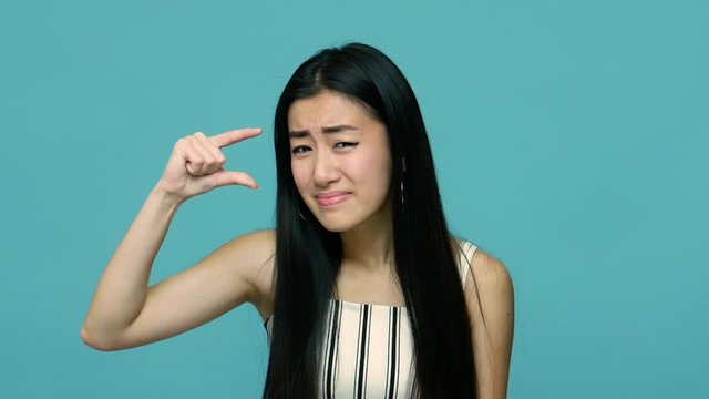 Positive asian woman with long black hair making little bit gesture, measuring small size and looking at camera with disappointed expression, showing minimum. studio shot isolated on blue background