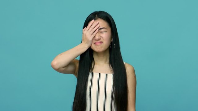 Oh no, i forgot! Unhappy asian woman with long straight black hair feeling sorrow, doing facepalm gesture with hand on face, feeling regret about loss, failure. studio shot isolated on blue background